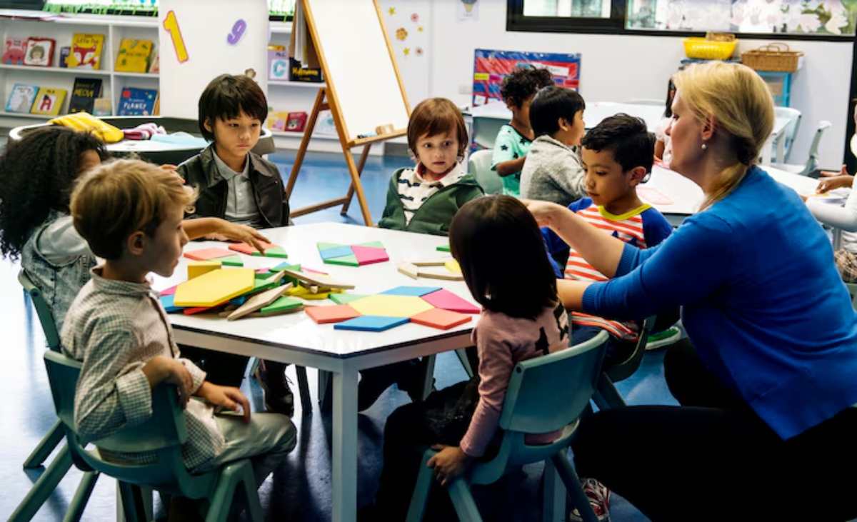 A teacher engages a small group of children at a table, surrounded by colorful geometric shapes and materials in a bright classroom.