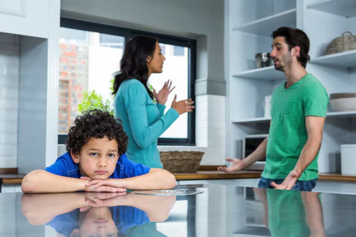 A young child sits at a kitchen island with a worried expression, while adults behind him appear to be arguing.