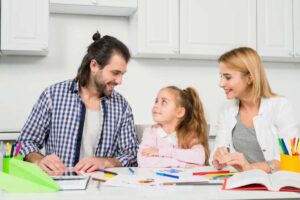 A family sits at a table covered with colorful art supplies and drawings, engaging in creative activities in a bright kitchen.