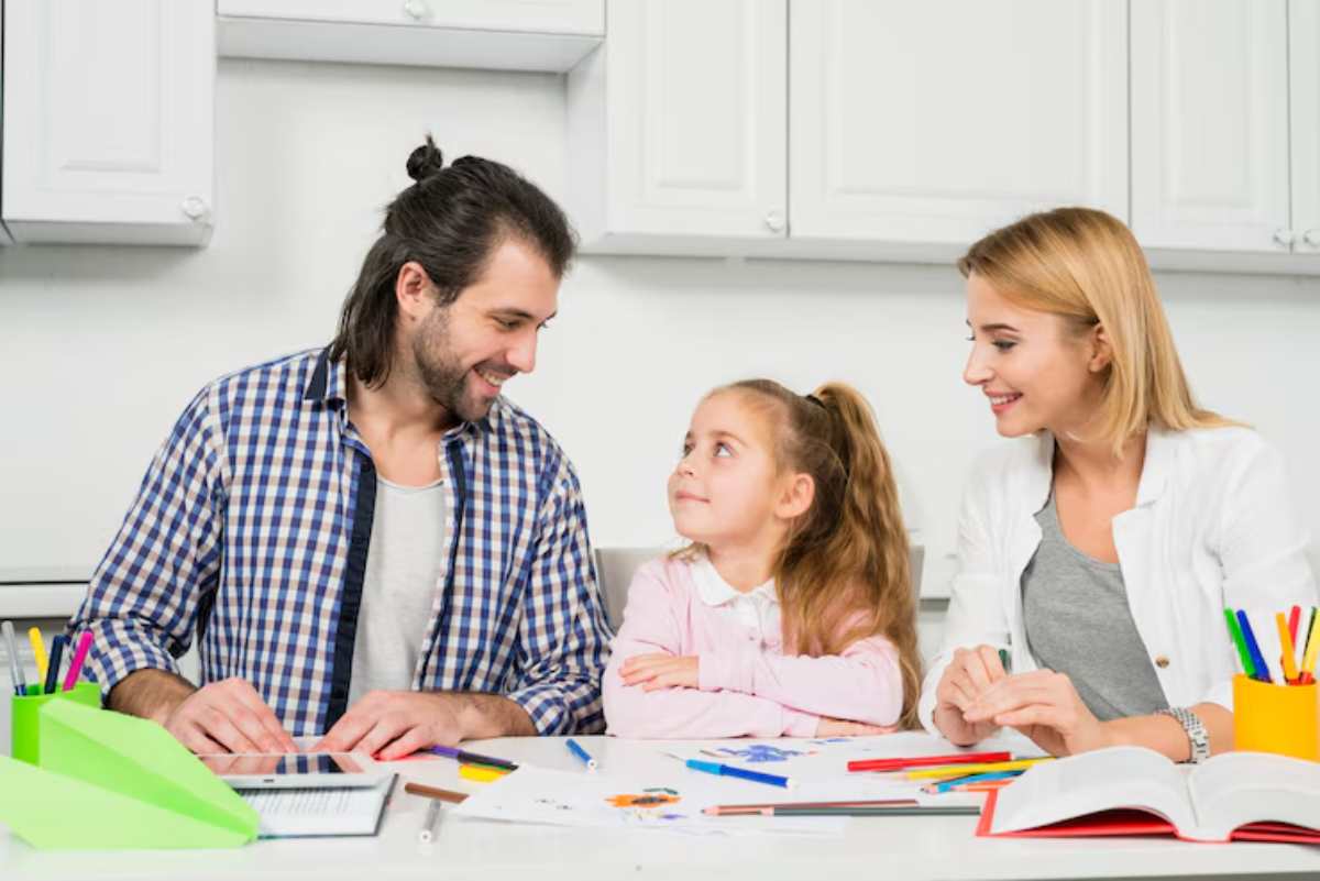 A family sits at a table covered with colorful art supplies and drawings, engaging in creative activities in a bright kitchen.