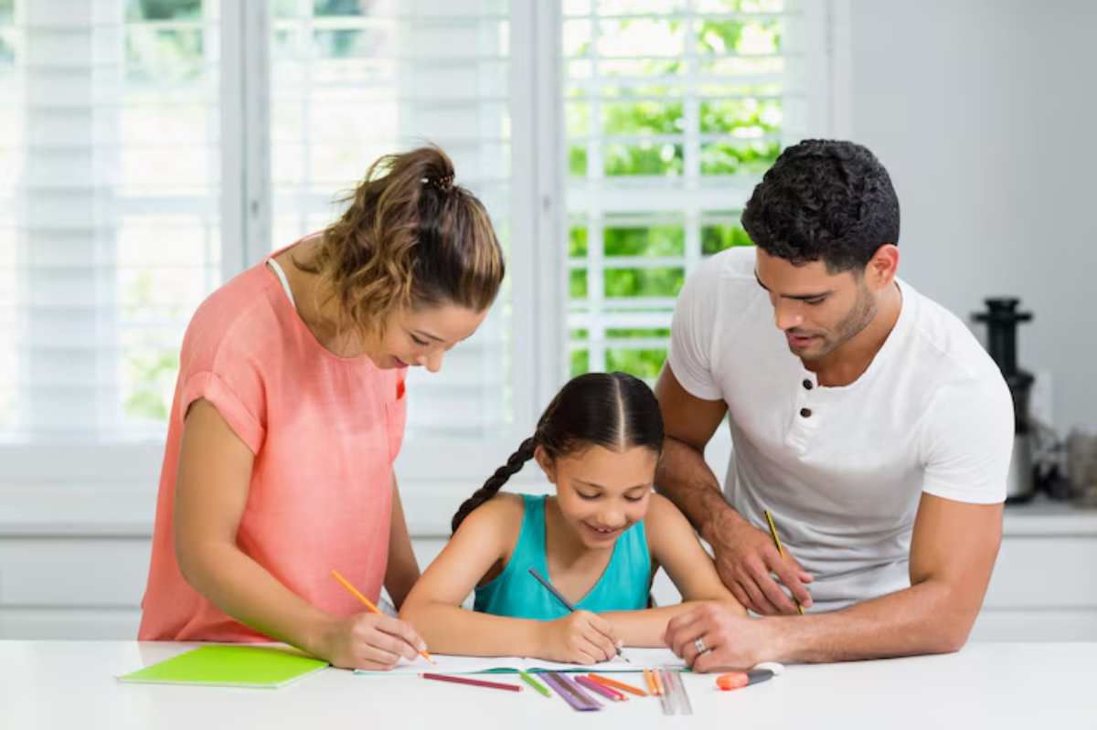 A child draws at a table, guided by parents, surrounded by colored pencils and a notebook in a bright, airy room with windows.