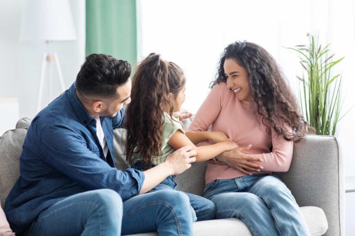 A man and woman sit on a couch, interacting with a young girl between them, in a well-lit living room with plants.