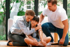 A woman and man kneel beside a young girl sitting on a wooden floor, providing comfort and support in a bright, inviting room.