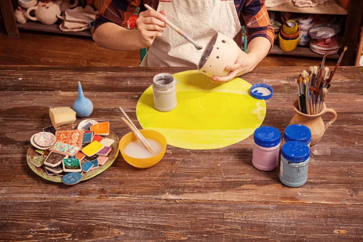 A person holding a paintbrush, decorating a ceramic pot, surrounded by pottery tools, paints, and colorful clay pieces on a wooden table.