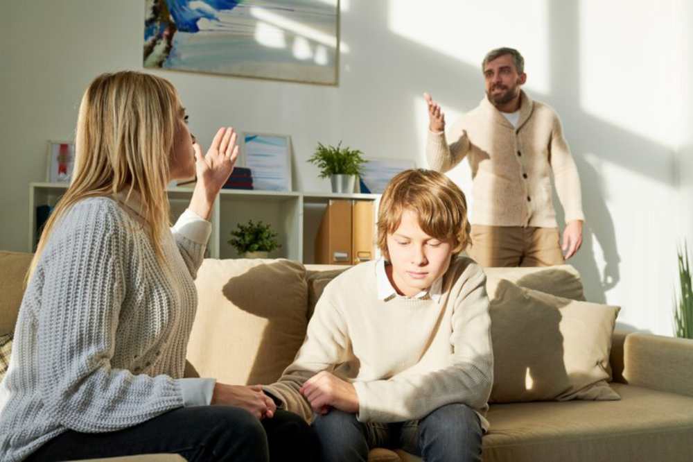 A tense family discussion takes place in a sunlit living room, with a woman gesturing and a child sitting silently on the sofa.