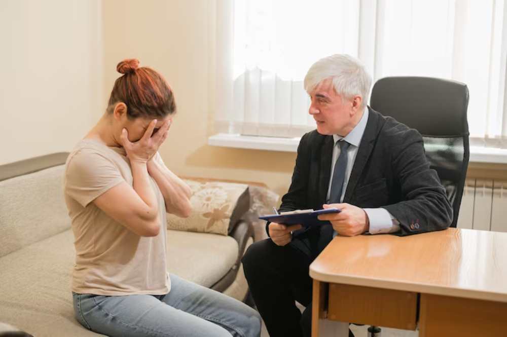 A seated woman appears distressed, while a man in a suit listens attentively, holding a clipboard in an office setting.