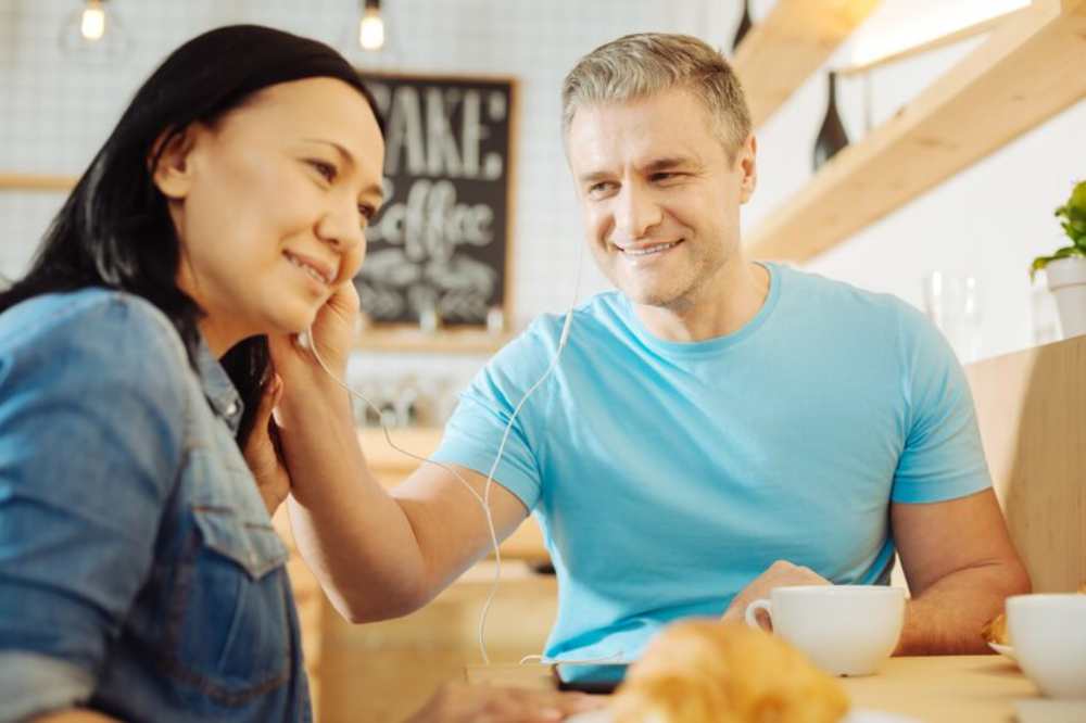 A couple sits at a table, sharing headphones while enjoying coffee and croissants in a cozy, warm atmosphere.