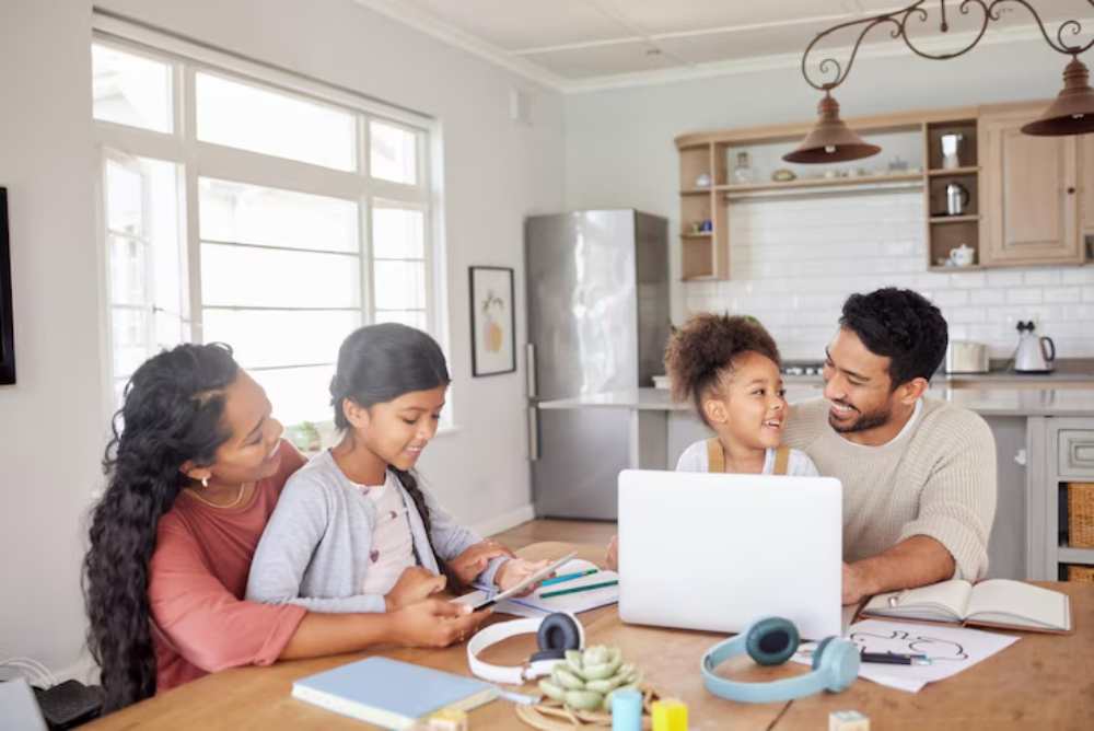 A group of family gather around a table with a laptop, books, and stationery in a bright, modern kitchen.