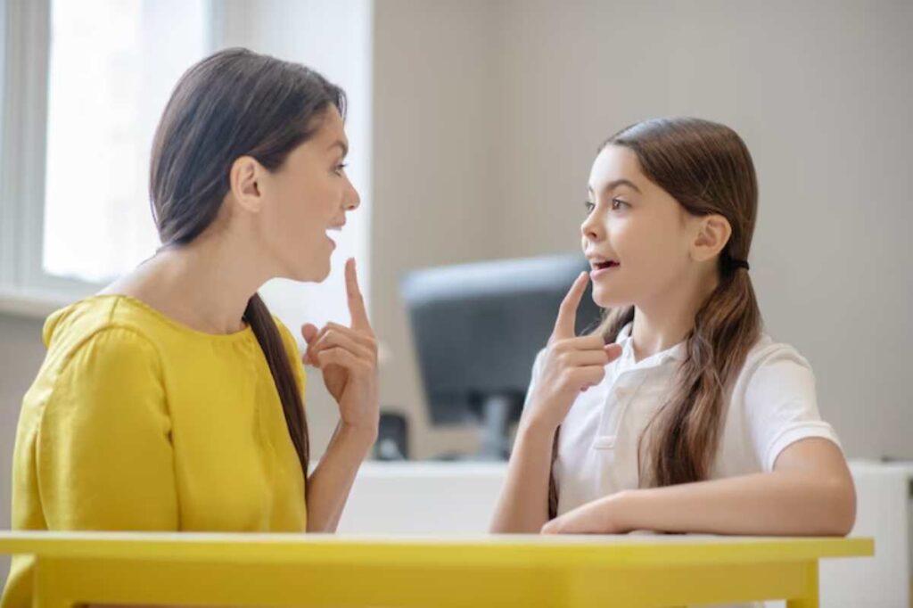 A woman and a child interact at a table, both thoughtfully gesturing and engaged in conversation in a bright, modern setting.