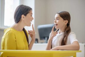 A woman and a child interact at a table, both thoughtfully gesturing and engaged in conversation in a bright, modern setting.
