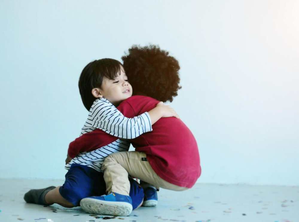 Two young children embrace each other while sitting on the floor, surrounded by scattered confetti against a light background.