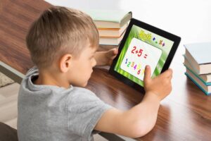 A child seated at a table, focused on a tablet displaying a math problem: '2+5='. Stacks of books are visible in the background.