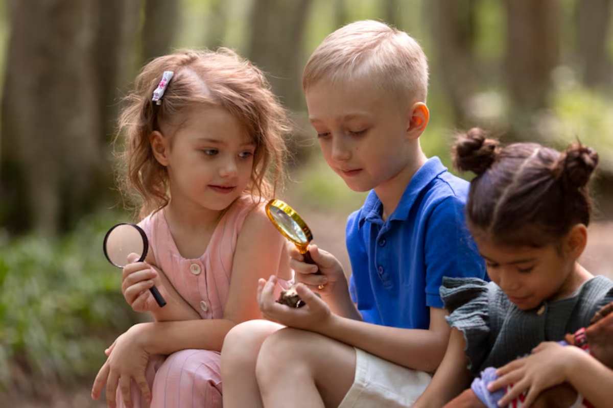 Three children sit together outdoors, holding magnifying glasses, exploring nature in a sunlit forest setting.