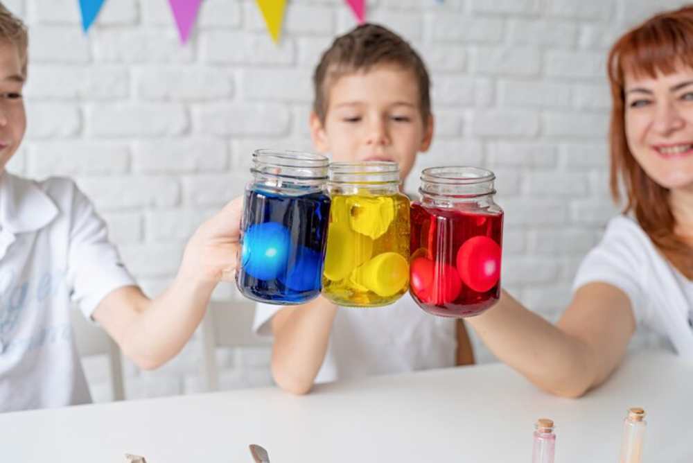 Three children hold jars of vibrant colored liquids — blue, yellow, and red — with colorful balls inside, celebrating a fun activity.