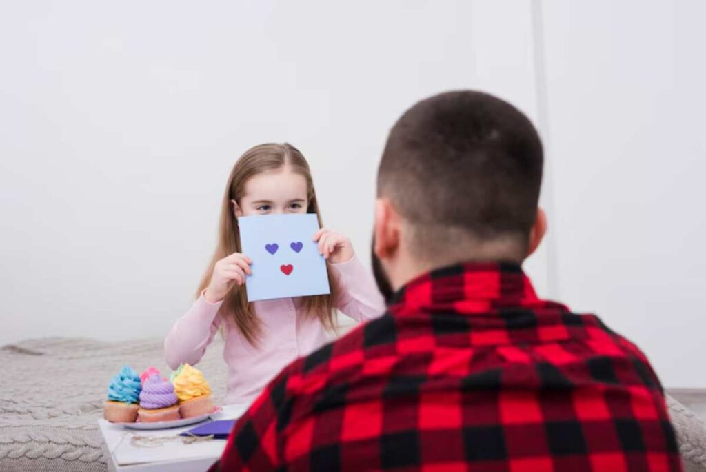 A child holds up a card with a red heart, while a person in a checkered shirt faces them, surrounded by colorful cupcakes on a table.