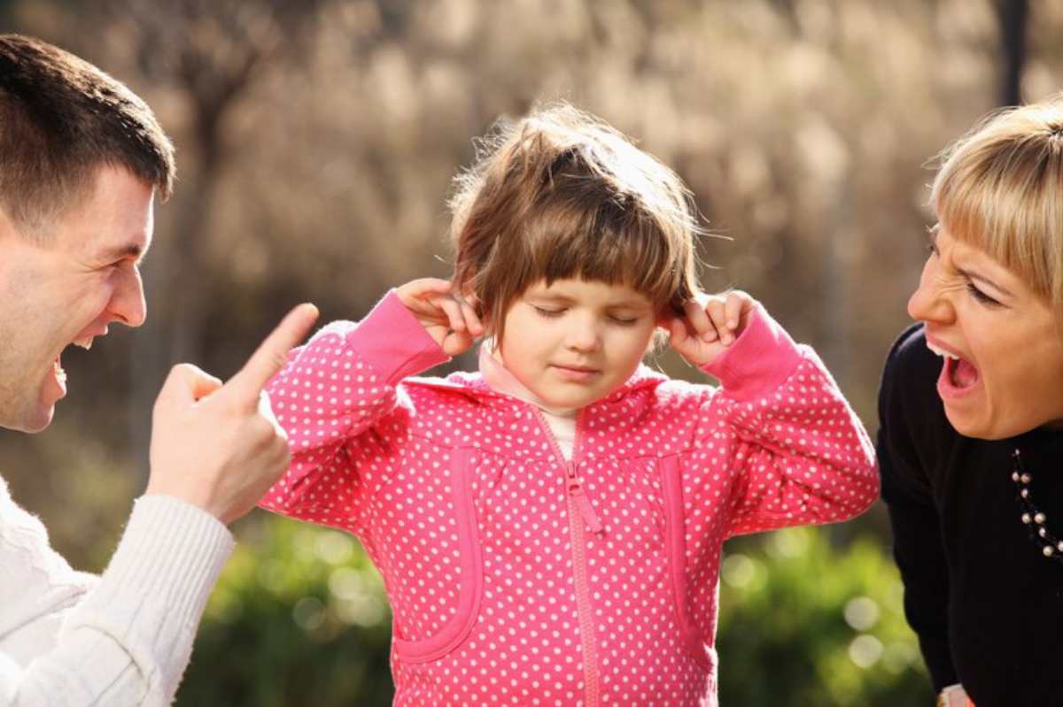 A child in a pink polka dot hoodie covers her ears while an adult gestures, set against a blurred outdoor background.
