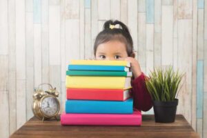 A child hides behind a stack of colorful books, with an alarm clock and a potted plant on a wooden table, against a wooden backdrop.