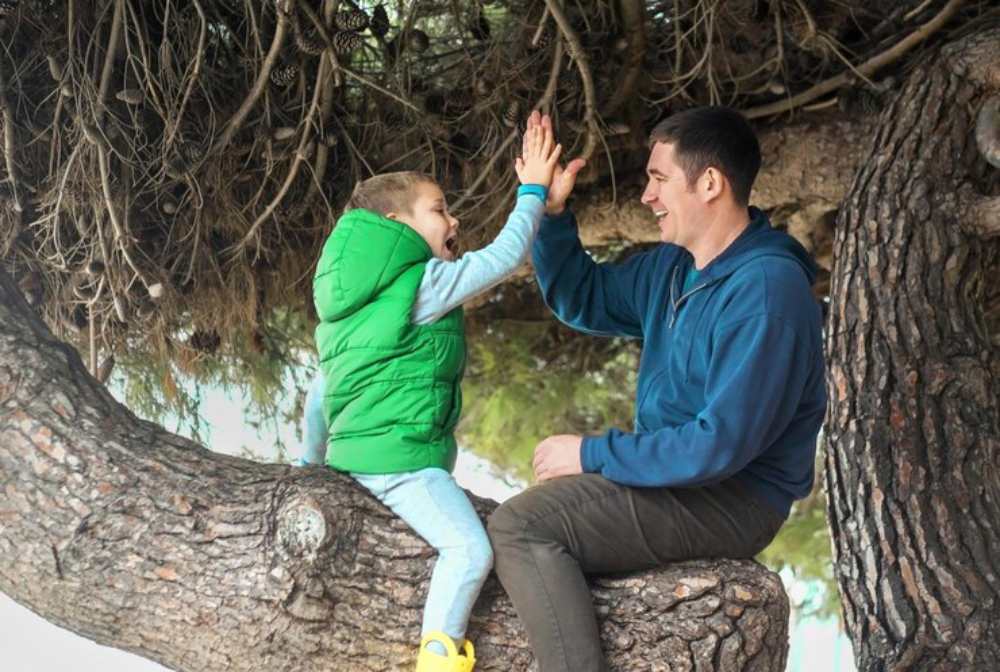 A child in a green vest sits on a tree branch, playfully touching hands with his father, surrounded by lush greenery.