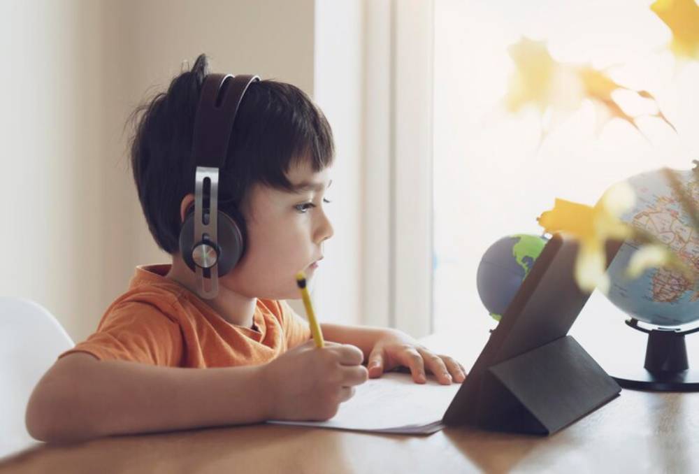 A child in an orange shirt wears headphones, writing notes at a desk with a tablet and a globe in a bright, cozy room.