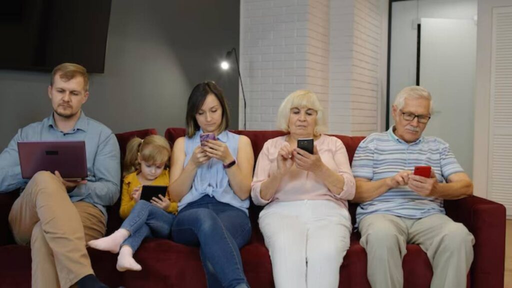 A family of five sitting on a maroon couch, each engaged with electronic devices, ranging from a laptop to smartphones.