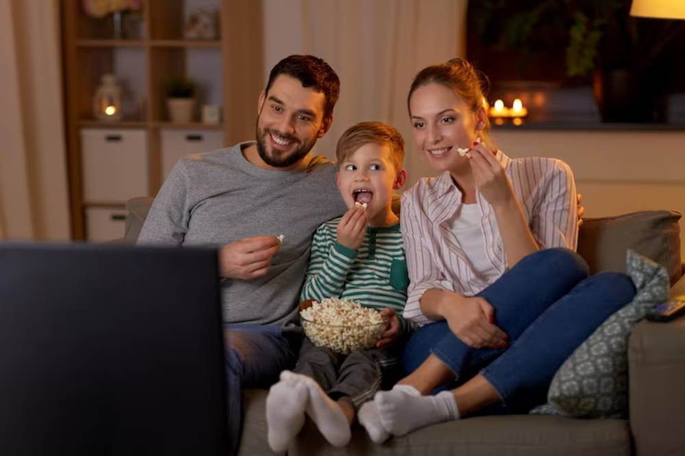 A family of three sits on a couch in a cozy living room, sharing popcorn while watching TV in a warm, ambient light.