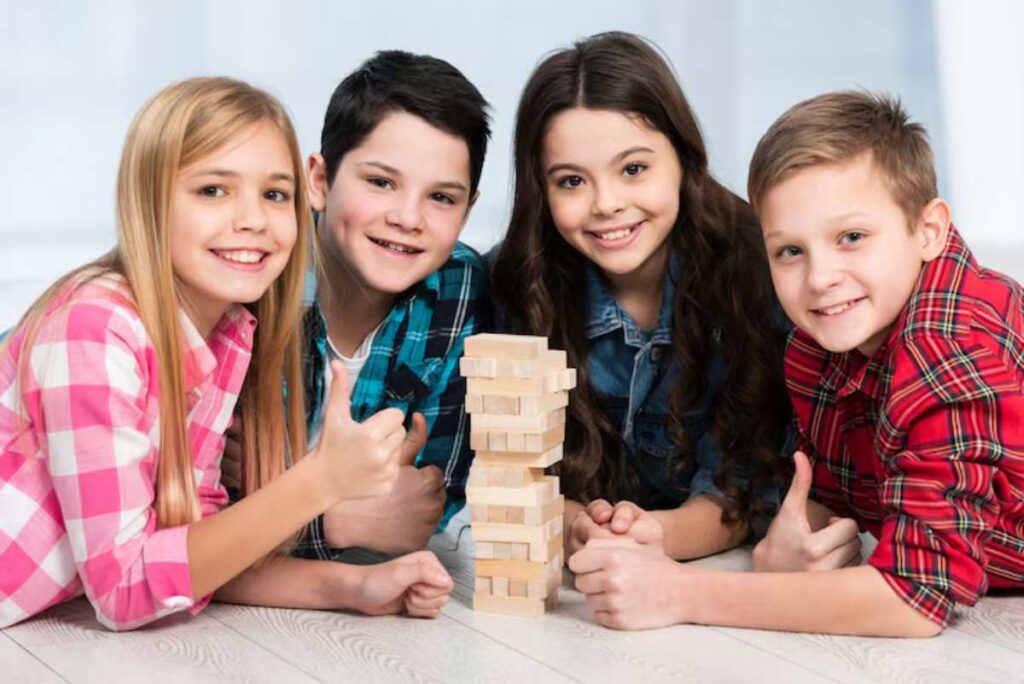 Four children gather around a Jenga tower, smiling and giving thumbs up, in a bright and cheerful indoor setting.