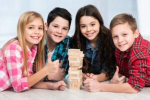 Four children gather around a Jenga tower, smiling and giving thumbs up, in a bright and cheerful indoor setting.
