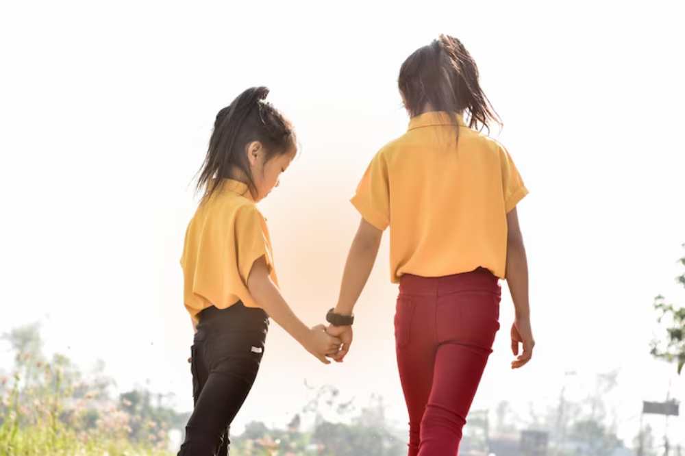 Two girls in yellow shirts and dark pants walk hand-in-hand in a scenic outdoor setting, illuminated by bright sunlight.