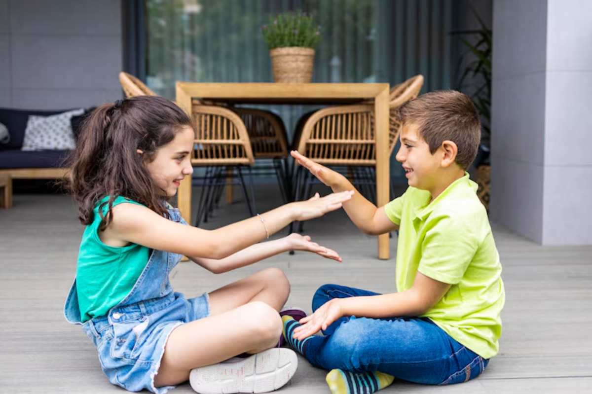 Two children sit on the floor of a modern patio, engaged in a playful hand game with smiles on their faces.