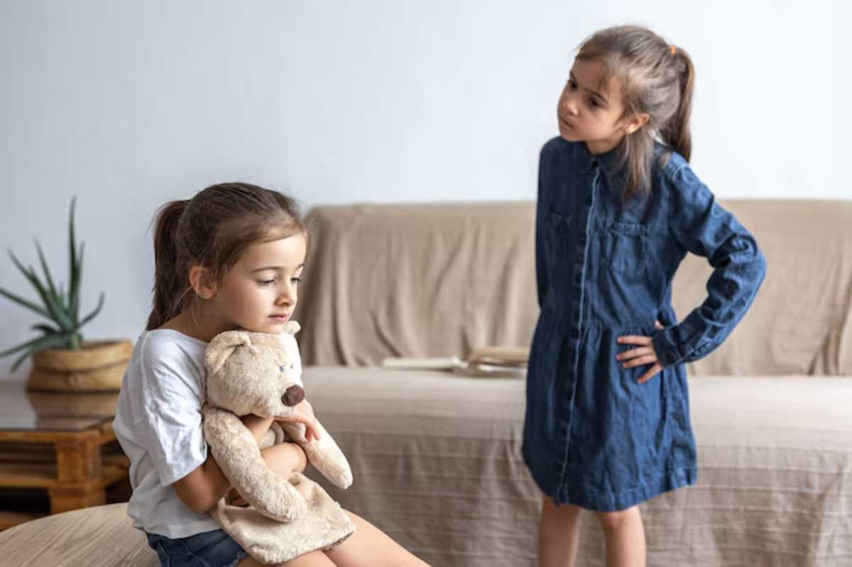 Two children interact indoors; one hugs a teddy bear while the other stands with hands on hips, both in casual clothing.