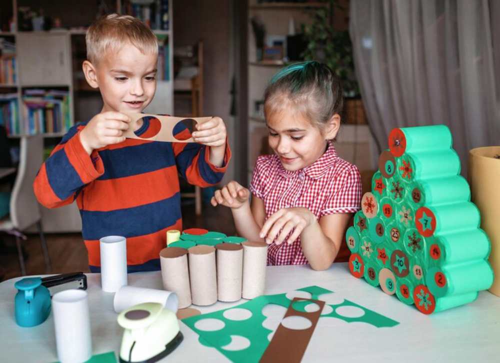 Two children are crafting at a table, surrounded by colorful paper tubes, craft supplies, and a green tree art project.