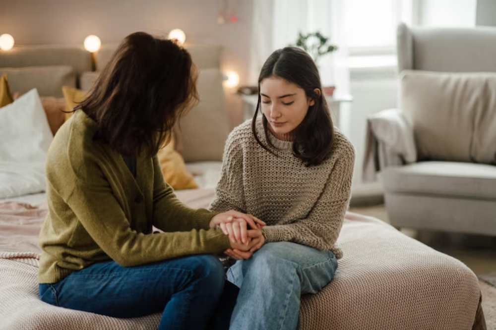 Two women sit closely on a bed, holding hands in a supportive manner, surrounded by a cozy, softly lit room adorned with cushions.