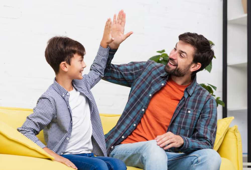 A teenager and an adult are sitting on a yellow couch, engaged in a playful high-five. A plant and a bookshelf are in the background.