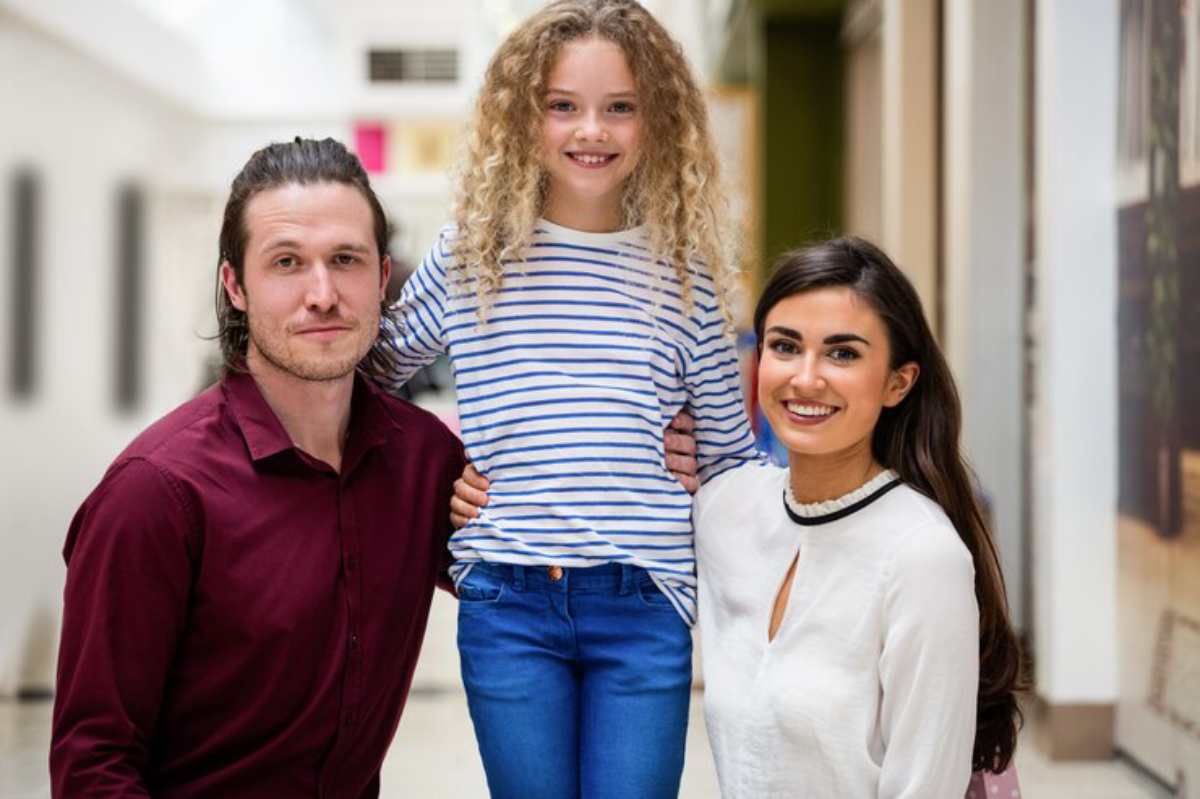 A smiling teenager wearing a striped shirt is held by a man in a red shirt and a woman in a white blouse, standing in a hallway.