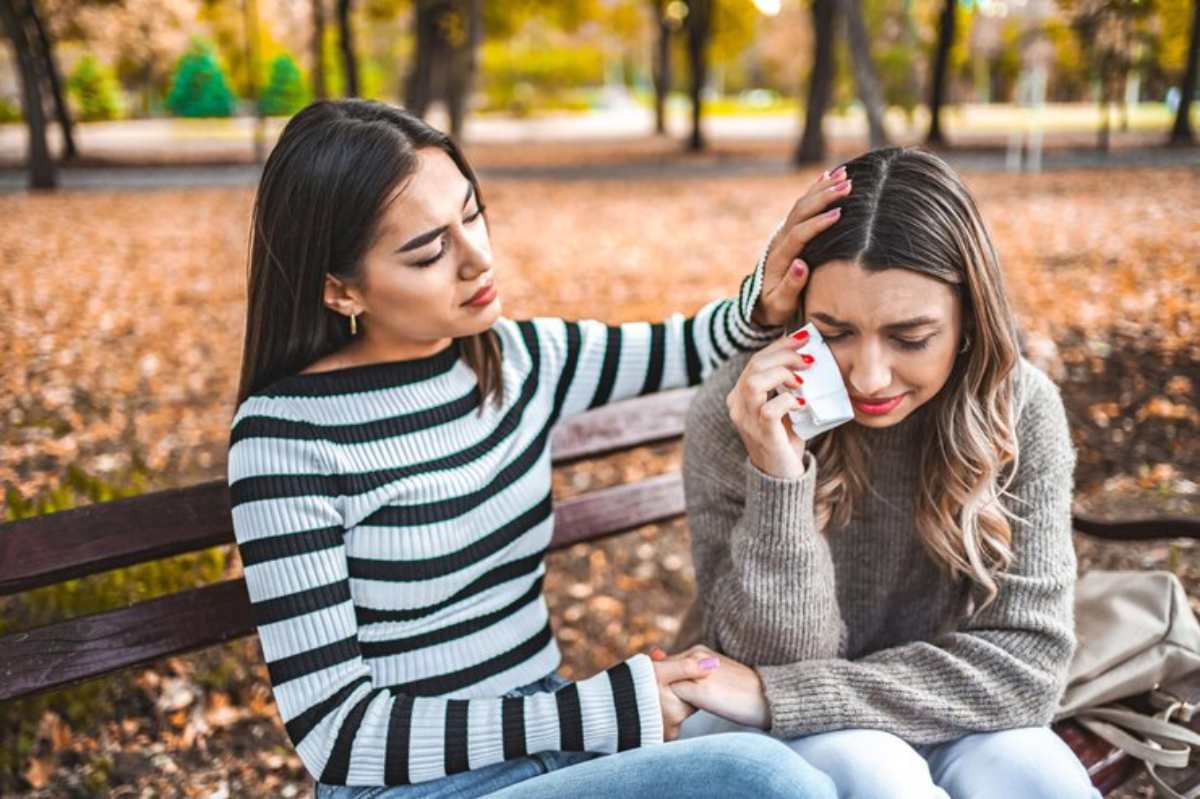 Two teenagers sit closely on a bench in a park during autumn, surrounded by fallen leaves, sharing a moment of connection and support.