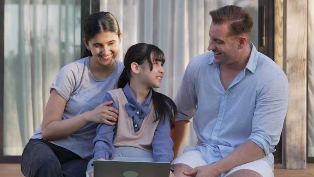 A happy family of three sitting outdoors, smiling at each other. The teen, in the center, holds a laptop. Warm, joyful atmosphere.