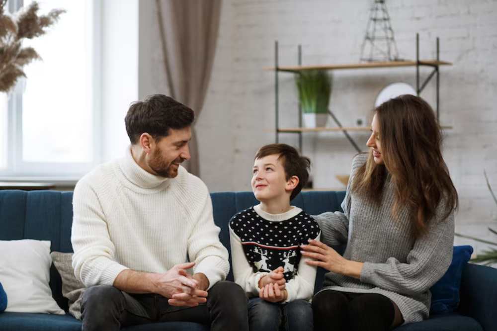 A family sits together on a blue couch, sharing a moment in a cozy living area with plants and decorative shelves in the background.