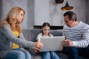 A woman and a man sit on a couch, sharing a laptop with a girl.