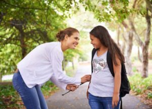 A woman adjusts a teenager's clothing in a park, surrounded by green trees and a pathway. The teenager carries a backpack.