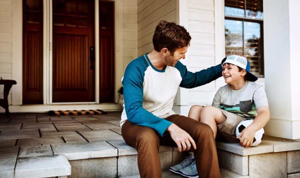 A man and a boy sit together on a porch step, sharing a warm conversation, with a soccer ball resting beside the boy.